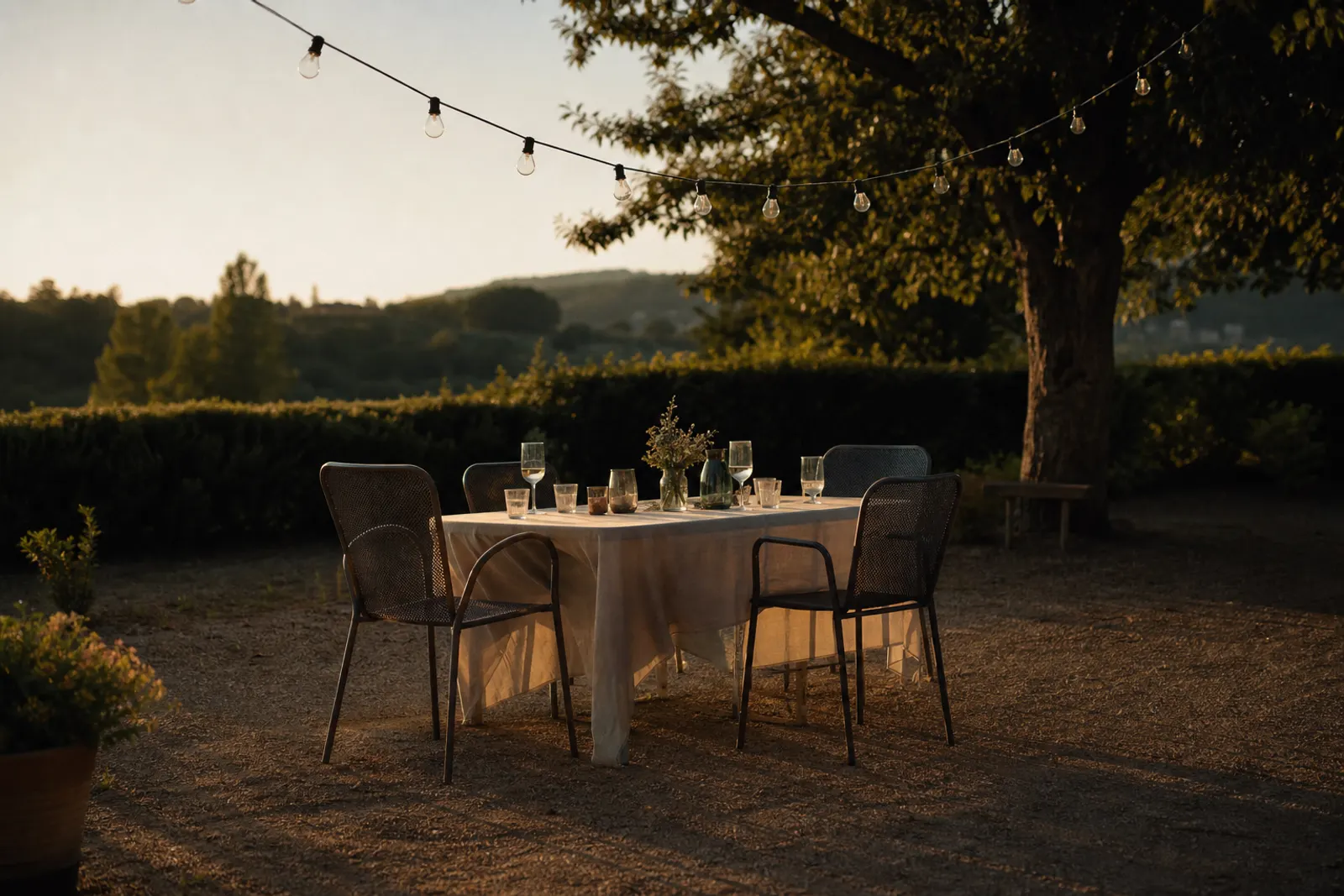 A garden table at dusk with four empty chairs and string lights overhead